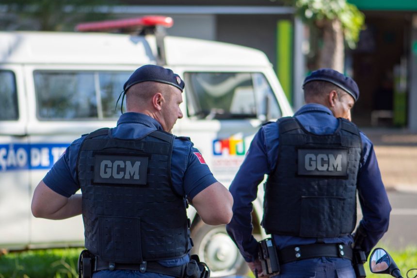 Dois guardas municipais uniformizados patrulham uma rua.