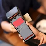 Close de uma mão aproximando um smartphone de um terminal para realizar um pagamento por NFC.