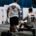 Competidor rema em um ergômetro enquanto é acompanhado por um juiz durante uma competição esportiva indoor.
