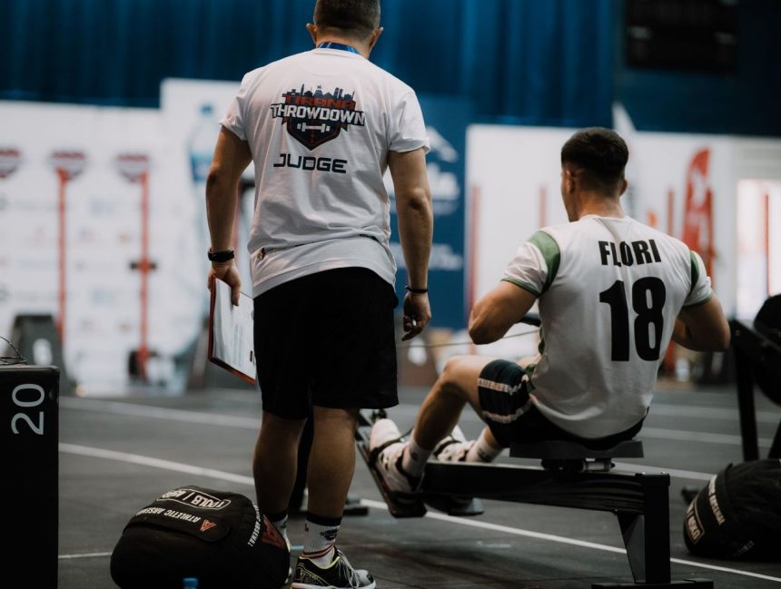 Competidor rema em um ergômetro enquanto é acompanhado por um juiz durante uma competição esportiva indoor.