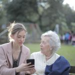 Mulher orienta uma idosa sentada em um banco de parque sobre o uso do celular.