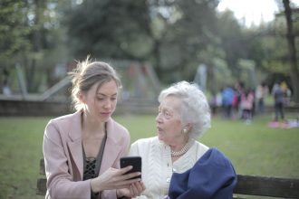 Mulher orienta uma idosa sentada em um banco de parque sobre o uso do celular.